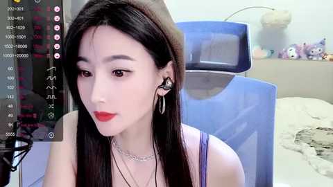 Media: Video of a young East Asian woman with fair skin, long black hair, and red lipstick, wearing a sleeveless purple top, sitting in a gaming chair, with a blue gaming desk and plush toys in the background.