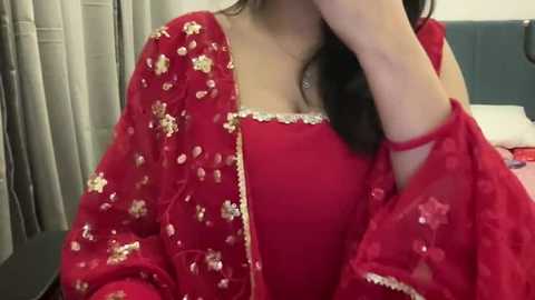 Media: Video of a woman in a red sari with gold floral embroidery, partially covered by a red dupatta with floral patterns. She has medium skin tone, long dark hair, and wears a red blouse. Background features a white curtain and a blue cushion.