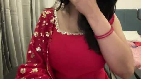 Video of a woman with medium skin tone, wearing a red sari with gold embroidery and a matching red blouse, her long dark hair cascading over her shoulder. Background shows a bed with pink sheets and grey curtains.