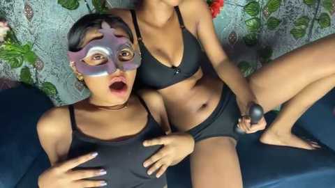 Media: Video of two women in black lingerie, one with a purple mask, both holding sex toys, on a blue couch, background with green leaves.