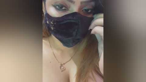 Media: Video of a woman with light skin, wearing a dark blue face mask with gold accents, green eye makeup, and a gold necklace, in a blurred, sepia-toned background.