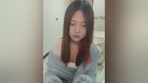 Media: A video of a young Asian woman with long, straight, dark brown hair, fair skin, and a soft, serene expression. She wears a light blue robe over a white top, sitting on a bed with a floral patterned blanket.