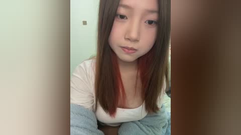 Media: A video of an Asian woman with long, straight, dark brown hair featuring red highlights. She has fair skin and wears a white top and light blue cardigan. The background is out of focus, with a plain wall.