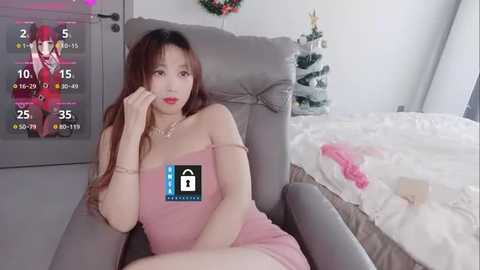 Media: A video of an East Asian woman in a pink dress, sitting in a gray chair, in a modern bedroom with a decorated Christmas tree and a bed with white sheets.