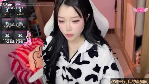 Media: Video of an East Asian woman with long black hair wearing a cow print hoodie, standing in a cozy room with pink walls and a plush toy.