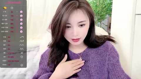 Media: A video of a young East Asian woman with long, dark hair, fair skin, and a purple sweater, sitting indoors with a potted plant in the background.