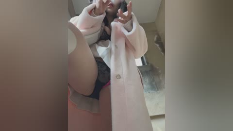 Media: Video of a slender, fair-skinned woman in black lace panties and a white bathrobe, striking a provocative pose with her legs spread, indoors with a beige wall and wooden floor.