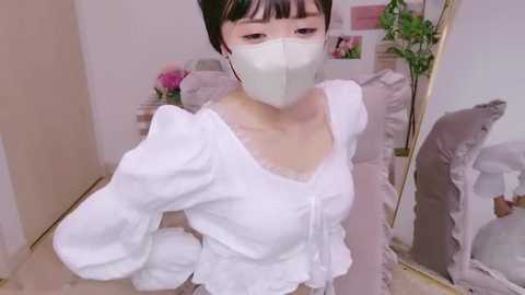 Media: A young Asian woman with short black hair, wearing a white mask, white blouse, and white pants, takes a selfie in a cozy, pink-tiled room with floral decor.