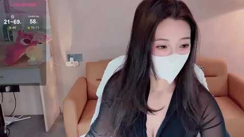 Video of an Asian woman with long black hair, wearing a white face mask and black jacket, sitting on a beige couch in a dimly lit room.