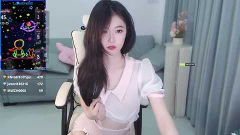 Media: Video of an Asian woman with long black hair, wearing a sheer white dress, sitting on a beige chair in a minimalist living room.