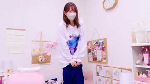 Media: Video of a young woman with straight brown hair, wearing a white hoodie with a blue graphic and blue pants, standing in a pastel-colored room with posters and stuffed toys.