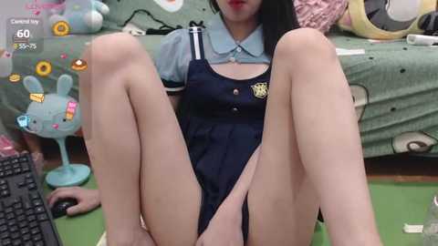 Media: Video of an Asian woman in a schoolgirl outfit, sitting on the floor with legs spread, wearing a blue dress with a yellow emblem, in a colorful, cluttered room.