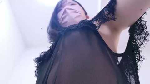 Media: Video of a woman with fair skin, medium-length brown hair, wearing a sheer black lace bra, lifting her arm, revealing her breasts. Background is blurred, white walls.