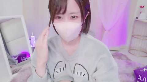 Media: A video of an East Asian woman with light skin, short brown hair, and a white surgical mask, wearing a grey \"SpongeBob\" sweatshirt, seated indoors with a white cat on a grey fur blanket.