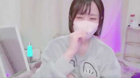 Video of an Asian woman with short dark hair, wearing a gray sweatshirt with cartoon eyes, covering her mouth with a white mask, in a cozy room with a pinkish-purple glow.