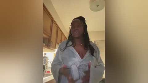 Video of a smiling Black woman with long braids, wearing a white bathrobe, standing in a kitchen with wooden cabinets and a white countertop.