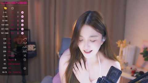 Media: Video of a smiling Asian woman with long brown hair, wearing headphones, in a dimly lit room with beige curtains and a desk.