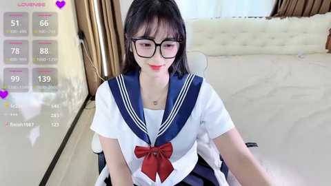 Media: Video of an East Asian woman with straight black hair, wearing a Japanese schoolgirl uniform, seated on a white bed with a beige headboard. She has fair skin, large-framed glasses, and a red bow.