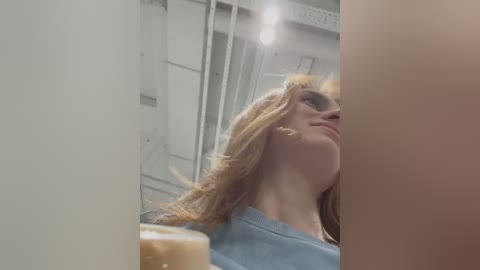 Media: A video captures a young woman with long, light brown hair, wearing a blue shirt, looking up thoughtfully. She is indoors, with a blurred, industrial background featuring white walls and a ceiling light.