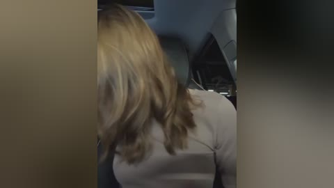 Media: Video of a woman with long, wavy blonde hair, wearing a light-colored jacket, seated in a car. The car's interior features gray seats and a steering wheel, creating a modern, clean look.