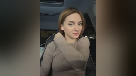 Media: Video of a Caucasian woman with shoulder-length auburn hair, wearing a light brown sweater, sitting in a car. She has a neutral expression and is seated in the driver's seat.