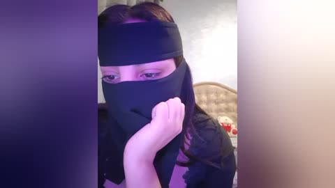 Media: A video of a young woman in a black ninja mask, with her hand covering her mouth, standing in a bedroom with a bed, teddy bear, and purple walls.