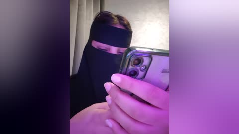 Media: Video of a person wearing a black blindfold, holding an iPhone, taking a selfie. The image is blurred with purple and pink hues.