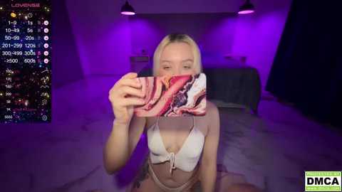 Media: A video of a blonde woman in a white bikini, kneeling on a bed in a dimly lit room, holding a phone to her face, with streaming stats displayed.