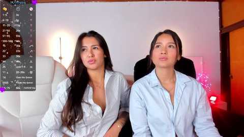 Media: Video of two young Asian women with long black hair, wearing light blue striped button-up shirts, sitting in an office with white furniture, pink lights, and a digital clock displaying 12:20.