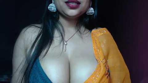 Media: Video of a South Asian woman with long dark hair, wearing a deep blue bra, a sheer orange sari with intricate embroidery, and large silver earrings. The background is dark and blurry, emphasizing her attire and jewelry.