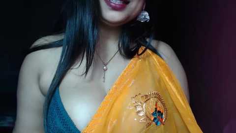 Media: Video of a South Asian woman with long black hair, wearing a blue sleeveless top, an orange sari with a gold emblem, and large silver earrings. She smiles slightly, her skin is light brown.