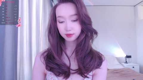 Media: Video of a young Asian woman with long, wavy brown hair, fair skin, and a slender physique, wearing a floral top. She stands in a minimalist bedroom with white walls, a bed, and a lamp.