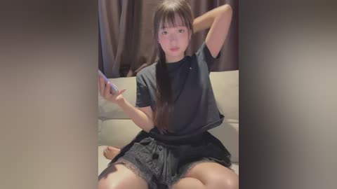 Media: Video of a young Asian woman with long, straight black hair and bangs, wearing a black crop top and shorts, sitting on a beige couch, holding a smartphone, in a dimly lit room with brown curtains.