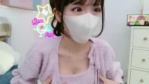 Media: Video of an Asian woman in a light pink, fluffy bathrobe with a white face mask, wearing a pink star-patterned headband, in a cozy bedroom setting.