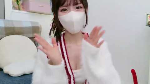 Media: Video of an Asian woman with fair skin, brown hair, wearing a white face mask, white robe, and red scarf, waving hands, in a modern, minimalist room with white walls and a beige sofa.