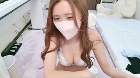 Media: Video of a young East Asian woman with long brown hair, wearing a white surgical mask, light pink bra, and sitting on a white rug in a cluttered bedroom with a bed and stuffed animals.