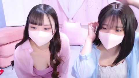 Media: Video of two young East Asian women with long black hair, wearing light pink face masks, pastel-colored tops, and sitting in a pink room.