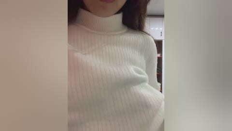 Video of a woman in a white ribbed turtleneck sweater, showing cleavage and part of her face. Background is blurry, featuring a white wall and a hint of a brick pattern.