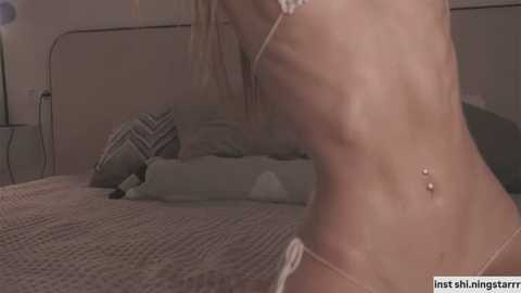 Media: A video of a slender, fair-skinned woman with blonde hair in a skimpy white bikini, standing beside a bed with gray bedding and pillows.