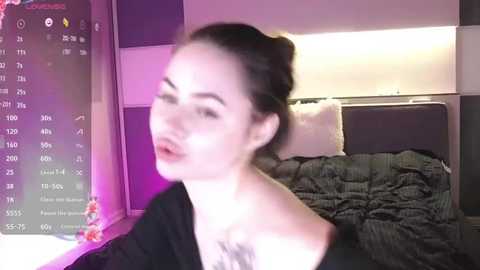 Media: Video of a fair-skinned woman with dark hair in a messy bun, wearing a black top, in a dimly lit bedroom with a messy bed and a digital clock displaying various time zones.