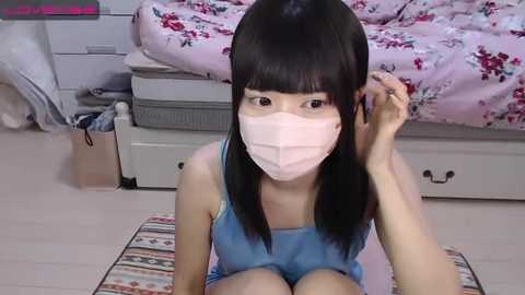 Media: Video of an East Asian woman with long black hair, wearing a pink face mask, blue tank top, and shorts, sitting on a striped rug in a pastel bedroom with a floral bedspread and white drawers.