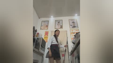 Media: Video of an Asian woman in a white kimono and black shorts, standing in a small, cluttered kitchen with three framed posters on the wall and a microwave and fridge visible.