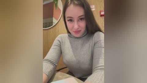 Media: Video of a young woman with long brown hair, light skin, and a slim build, wearing a gray ribbed sweater, seated at a table. Background shows a wooden wall with a circular mirror and a red fire alarm.