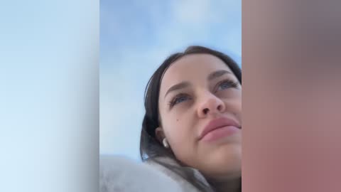 Media: Video of a young woman with light skin, blue eyes, and dark hair, gazing upward. She wears a white sweater and sits against a light blue backdrop.