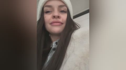 Media: Video of a young woman with long, dark hair, wearing a white beanie and a light-colored coat, standing in a dimly lit hallway.