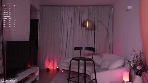 Media: Video of a modern living room with a white sectional sofa, black metal stool, and a tall, curved floor lamp. Soft, ambient lighting casts a pink hue, with red candles creating a cozy atmosphere.