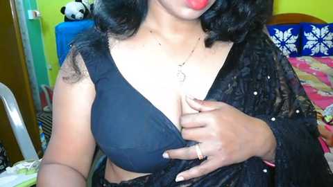 Media: Video of a South Asian woman with medium skin tone, black hair, wearing a black lace shawl over a black bra, exposing cleavage, and a gold necklace with a pendant. Background includes a bed with floral pillows and a stuffed panda.