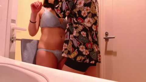 A video of a woman in light blue lingerie, standing in a bathroom, holding a colorful floral shirt. The background features a white door and a blue towel hanging on a hook.