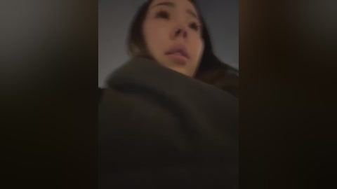 Media: A blurred, close-up video of a young woman with fair skin, long dark hair, and a slightly open mouth, wrapped in a dark, thick blanket, against a dimly lit, dark background.