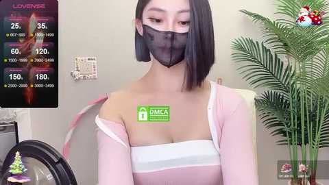 Media: Video of an Asian woman with straight black hair, wearing a black face mask and a light pink off-shoulder top, sitting in a room with a green plant and digital clock.